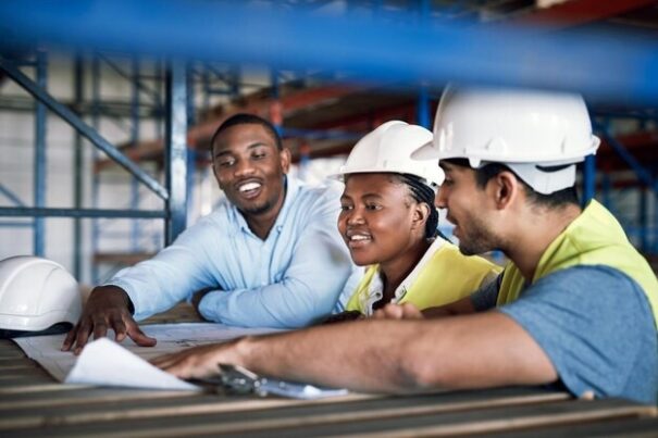 Three construction workers, wearing hard hats, discussing plans at a worksite.
